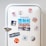 A white vintage refrigerator adorned with various colorful magnets and photos. There are images of a cityscape, a couple standing together, a heart-shaped object, a plate of food, and a circular magnet with an orange theme. The word 'FAMOUS' is spelled out using cut-up letters on separate magnets. Additional round magnets include a pattern of colorful stripes and sliced grapefruits.