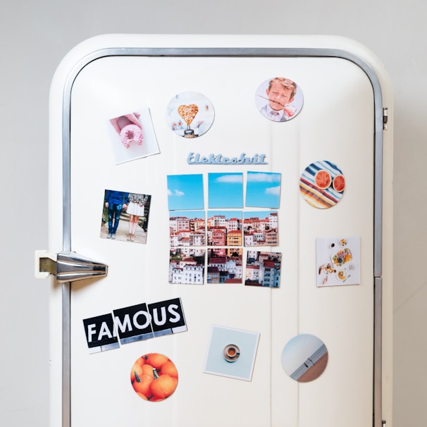 A white vintage refrigerator adorned with various colorful magnets and photos. There are images of a cityscape, a couple standing together, a heart-shaped object, a plate of food, and a circular magnet with an orange theme. The word 'FAMOUS' is spelled out using cut-up letters on separate magnets. Additional round magnets include a pattern of colorful stripes and sliced grapefruits.
