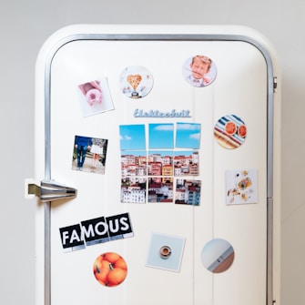 A white vintage refrigerator adorned with various colorful magnets and photos. There are images of a cityscape, a couple standing together, a heart-shaped object, a plate of food, and a circular magnet with an orange theme. The word 'FAMOUS' is spelled out using cut-up letters on separate magnets. Additional round magnets include a pattern of colorful stripes and sliced grapefruits.