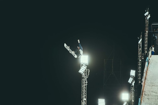 A snowboarder is performing an aerial trick high in the air at night, illuminated by bright floodlights. The scene captures the dynamic movement against a stark dark sky with the snowboarder's body twisting mid-air. Metal scaffolding and lights provide an industrial backdrop, adding to the extreme sports atmosphere.
