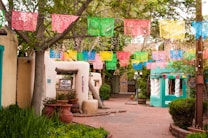 A quaint, colorful street adorned with vibrant papel picado banners hanging above. The adobe-style buildings feature rustic wooden doors and windows. There are potted plants and greenery surrounding the pathway, adding to the lively atmosphere.