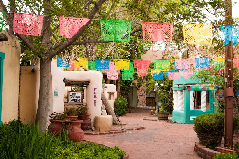 A quaint, colorful street adorned with vibrant papel picado banners hanging above. The adobe-style buildings feature rustic wooden doors and windows. There are potted plants and greenery surrounding the pathway, adding to the lively atmosphere.