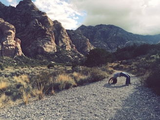A person is performing a backbend on a rocky path surrounded by a desert landscape. Rugged, towering mountains form the backdrop under a partly cloudy sky. Sparse vegetation and shrubs are scattered around the area.