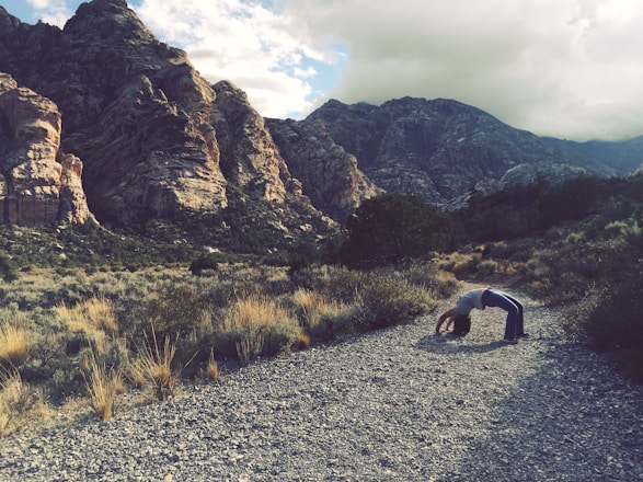 A person is performing a backbend on a rocky path surrounded by a desert landscape. Rugged, towering mountains form the backdrop under a partly cloudy sky. Sparse vegetation and shrubs are scattered around the area.