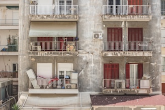 A weathered apartment building with visible signs of wear and age. Multiple balconies feature various objects like an air conditioning unit, chairs, and miscellaneous items. The building’s facade is gray with peeling paint, and the shutters on the windows are painted bright red. The scene suggests a sense of disrepair but also daily life with personal belongings scattered.