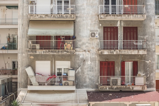 A weathered apartment building with visible signs of wear and age. Multiple balconies feature various objects like an air conditioning unit, chairs, and miscellaneous items. The building’s facade is gray with peeling paint, and the shutters on the windows are painted bright red. The scene suggests a sense of disrepair but also daily life with personal belongings scattered.