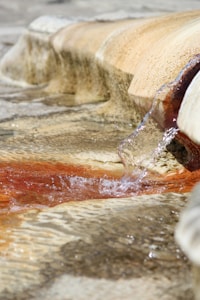 Clear water is flowing over textured stone surfaces forming gentle ripples. Some of the stone surfaces appear to be stained with a rust-colored discoloration, possibly from iron deposits or algae. The water is flowing smoothly, creating a calm and soothing atmosphere.