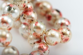 A close-up view of a strand of glossy pearls. Each pearl has a unique marbled design with shades of red, white, and subtle hints of green. The background is softly blurred, emphasizing the intricate patterns on the pearls.