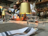 A restaurant bar setting with a bartender focused on pouring a drink from a large container. The counter is elegantly set with glasses, a napkin with a fork, and a small decorative plant. The background features shelves lined with bottles of various liquors, dimly lit by stylish tabletop lamps.