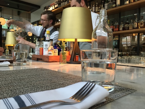 A restaurant bar setting with a bartender focused on pouring a drink from a large container. The counter is elegantly set with glasses, a napkin with a fork, and a small decorative plant. The background features shelves lined with bottles of various liquors, dimly lit by stylish tabletop lamps.