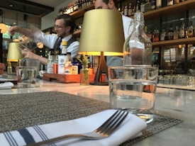 A restaurant bar setting with a bartender focused on pouring a drink from a large container. The counter is elegantly set with glasses, a napkin with a fork, and a small decorative plant. The background features shelves lined with bottles of various liquors, dimly lit by stylish tabletop lamps.