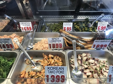 A display case filled with a variety of prepared seafood dishes, each labeled with a name and price. The selections include poke with labels like Limukohu Ahi Poke and Korean Tako Poke, along with other items such as Spicy Lobster, Soybeans with Roasted Garlic, and Kimchee Cucumber. The dishes are arranged in metal trays with serving utensils, on a refrigerated counter.