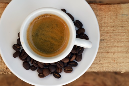 A cup of espresso rests on a white saucer scattered with coffee beans, set on a wooden surface.