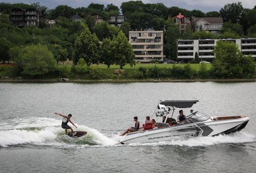 A speedboat is pulling a person wakeboarding on a river or lake. The wakeboarder is riding the waves with a stance that suggests skill and balance. Several people are on the boat, either watching or assisting the activity. The background features a line of trees and modern residential buildings.