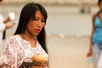 A woman is holding a musical instrument labeled 'SIGU' while standing outdoors. She is dressed in a patterned blouse and appears thoughtful or engaged in the activity. The background is blurred, showing a beach or natural setting with other indistinct figures around.
