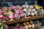 An assortment of small flower bouquets in various colors are arranged neatly on wooden shelves. The bouquets feature bright and vibrant flowers such as daisies, roses, and carnations. The display has signage with pricing details in a retail setting.