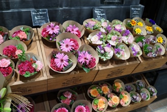 An assortment of small flower bouquets in various colors are arranged neatly on wooden shelves. The bouquets feature bright and vibrant flowers such as daisies, roses, and carnations. The display has signage with pricing details in a retail setting.