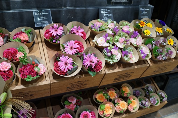 An assortment of small flower bouquets in various colors are arranged neatly on wooden shelves. The bouquets feature bright and vibrant flowers such as daisies, roses, and carnations. The display has signage with pricing details in a retail setting.