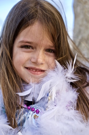 A young girl with long brown hair is smiling while adorned in a feather boa. A necklace with purple beads is around her neck, adding a playful and colorful touch.