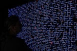 A large digital screen displays a complex pattern resembling a maze or circuit board. The design is composed of bright blue lines and dots, interspersed with small red lights scattered throughout. In the foreground, a silhouette of a person is visible, observing the intricate display.