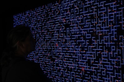 A large digital screen displays a complex pattern resembling a maze or circuit board. The design is composed of bright blue lines and dots, interspersed with small red lights scattered throughout. In the foreground, a silhouette of a person is visible, observing the intricate display.
