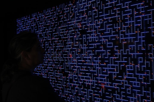 A large digital screen displays a complex pattern resembling a maze or circuit board. The design is composed of bright blue lines and dots, interspersed with small red lights scattered throughout. In the foreground, a silhouette of a person is visible, observing the intricate display.
