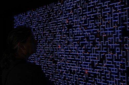 A large digital screen displays a complex pattern resembling a maze or circuit board. The design is composed of bright blue lines and dots, interspersed with small red lights scattered throughout. In the foreground, a silhouette of a person is visible, observing the intricate display.