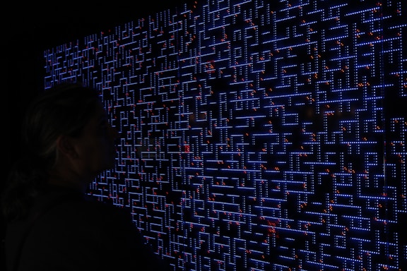 A large digital screen displays a complex pattern resembling a maze or circuit board. The design is composed of bright blue lines and dots, interspersed with small red lights scattered throughout. In the foreground, a silhouette of a person is visible, observing the intricate display.