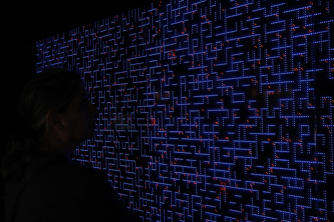 A large digital screen displays a complex pattern resembling a maze or circuit board. The design is composed of bright blue lines and dots, interspersed with small red lights scattered throughout. In the foreground, a silhouette of a person is visible, observing the intricate display.