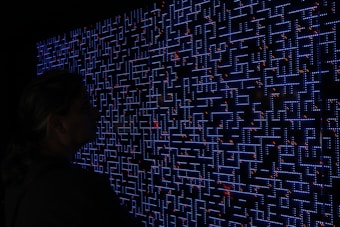 A large digital screen displays a complex pattern resembling a maze or circuit board. The design is composed of bright blue lines and dots, interspersed with small red lights scattered throughout. In the foreground, a silhouette of a person is visible, observing the intricate display.