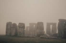 A foggy landscape featuring a prehistoric stone circle with large standing stones arranged in a circular formation. The grass in the foreground appears moist and muted due to the fog, which envelops the entire scene, imparting an air of mystery and antiquity.