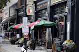 A quaint sidewalk scene features outdoor seating with green umbrellas in front of a brick-and-mortar business. Signs for parking regulations are prominently displayed. Flower pots add color to the scene, which includes various storefronts with vintage signage and clothing displays.