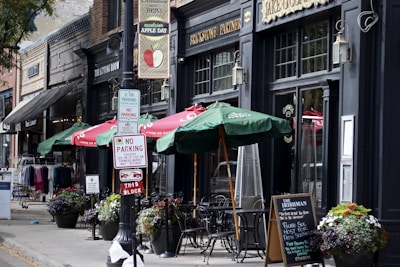 A quaint sidewalk scene features outdoor seating with green umbrellas in front of a brick-and-mortar business. Signs for parking regulations are prominently displayed. Flower pots add color to the scene, which includes various storefronts with vintage signage and clothing displays.
