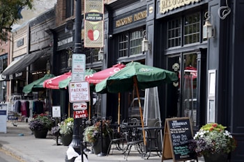 A quaint sidewalk scene features outdoor seating with green umbrellas in front of a brick-and-mortar business. Signs for parking regulations are prominently displayed. Flower pots add color to the scene, which includes various storefronts with vintage signage and clothing displays.