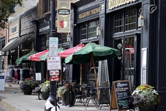 A quaint sidewalk scene features outdoor seating with green umbrellas in front of a brick-and-mortar business. Signs for parking regulations are prominently displayed. Flower pots add color to the scene, which includes various storefronts with vintage signage and clothing displays.