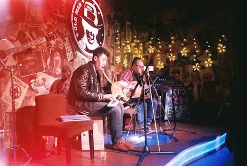 Two musicians with guitars perform on a small stage adorned with eclectic decorations and fairy lights. The room is dimly lit, creating a cozy, intimate atmosphere. A red chair and open notebook are visible, while posters and various artwork cover the walls.