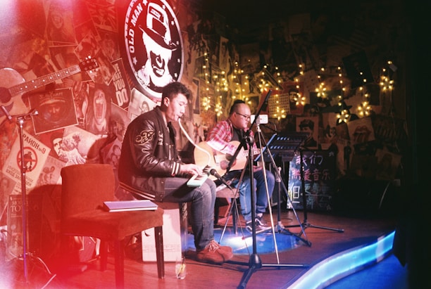 Two musicians with guitars perform on a small stage adorned with eclectic decorations and fairy lights. The room is dimly lit, creating a cozy, intimate atmosphere. A red chair and open notebook are visible, while posters and various artwork cover the walls.