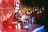 Two musicians with guitars perform on a small stage adorned with eclectic decorations and fairy lights. The room is dimly lit, creating a cozy, intimate atmosphere. A red chair and open notebook are visible, while posters and various artwork cover the walls.