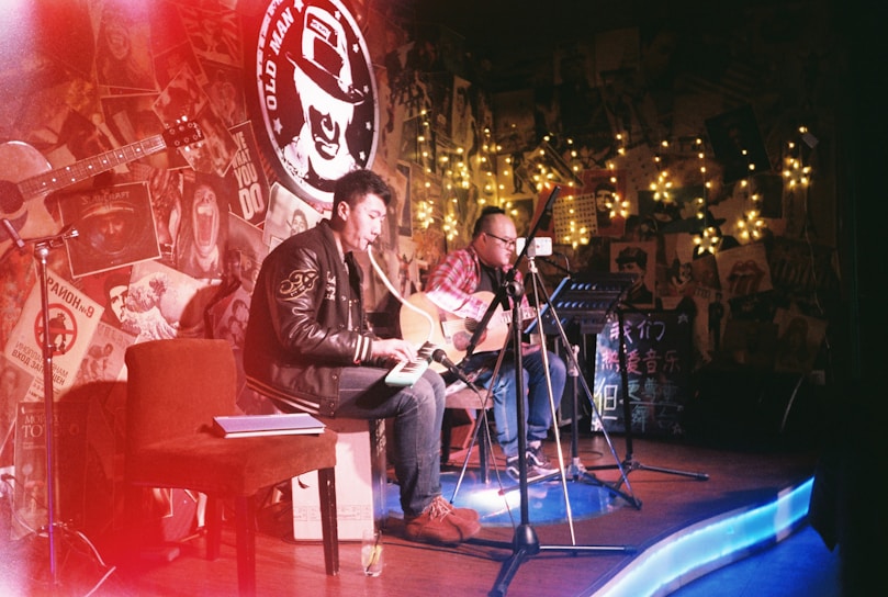 Two musicians with guitars perform on a small stage adorned with eclectic decorations and fairy lights. The room is dimly lit, creating a cozy, intimate atmosphere. A red chair and open notebook are visible, while posters and various artwork cover the walls.