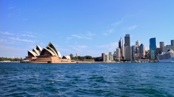The image presents a waterside view of a prominent opera house with unique architectural design alongside a city skyline featuring modern skyscrapers under a clear blue sky. A large cruise ship is docked nearby.