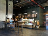 A cozy coffee shop interior with modern decor features a counter with illuminated display cases filled with drinks and pastries. A menu board hangs above the counter, and industrial-style pendant lights provide warm lighting. Two children are sitting on high stools at a long wooden table, engaged with their surroundings.