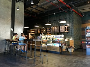 A cozy coffee shop interior with modern decor features a counter with illuminated display cases filled with drinks and pastries. A menu board hangs above the counter, and industrial-style pendant lights provide warm lighting. Two children are sitting on high stools at a long wooden table, engaged with their surroundings.