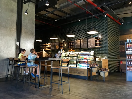 A cozy coffee shop interior with modern decor features a counter with illuminated display cases filled with drinks and pastries. A menu board hangs above the counter, and industrial-style pendant lights provide warm lighting. Two children are sitting on high stools at a long wooden table, engaged with their surroundings.
