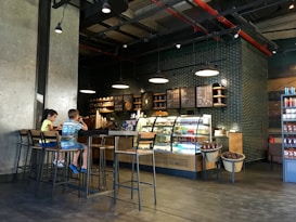 A cozy coffee shop interior with modern decor features a counter with illuminated display cases filled with drinks and pastries. A menu board hangs above the counter, and industrial-style pendant lights provide warm lighting. Two children are sitting on high stools at a long wooden table, engaged with their surroundings.