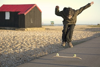 A person is performing a trick over a skateboard on a paved path near a beach. In the background, there is a small black wooden hut with a red roof. The surrounding landscape consists of a pebble-covered area and a view of the distant sea.
