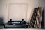 A vintage turntable is placed on a shelf alongside a collection of vinyl records stacked vertically. The turntable has a clear dust cover partially open, and the records have varying cover designs, suggesting a diverse collection. Soft lighting creates a nostalgic atmosphere.