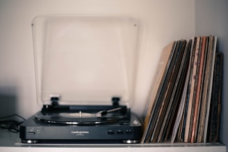 A vintage turntable is placed on a shelf alongside a collection of vinyl records stacked vertically. The turntable has a clear dust cover partially open, and the records have varying cover designs, suggesting a diverse collection. Soft lighting creates a nostalgic atmosphere.
