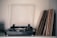 A vintage turntable is placed on a shelf alongside a collection of vinyl records stacked vertically. The turntable has a clear dust cover partially open, and the records have varying cover designs, suggesting a diverse collection. Soft lighting creates a nostalgic atmosphere.