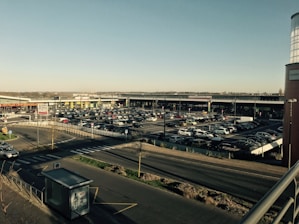 A shopping center parking lot filled with numerous cars under a clear blue sky. The area is expansive with rows of parked vehicles, roads, and a visible pedestrian crossing. There's a bus stop structure in the foreground and various retail stores in the background.