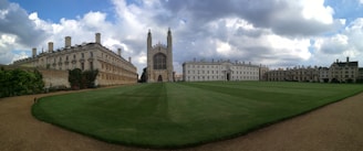 A large open grassy courtyard is bordered by historic stone buildings with numerous windows and chimneys. In the center, a prominent gothic-style chapel stands with intricate architecture and tall spires. The sky is partly cloudy, creating a dramatic backdrop for the scene.